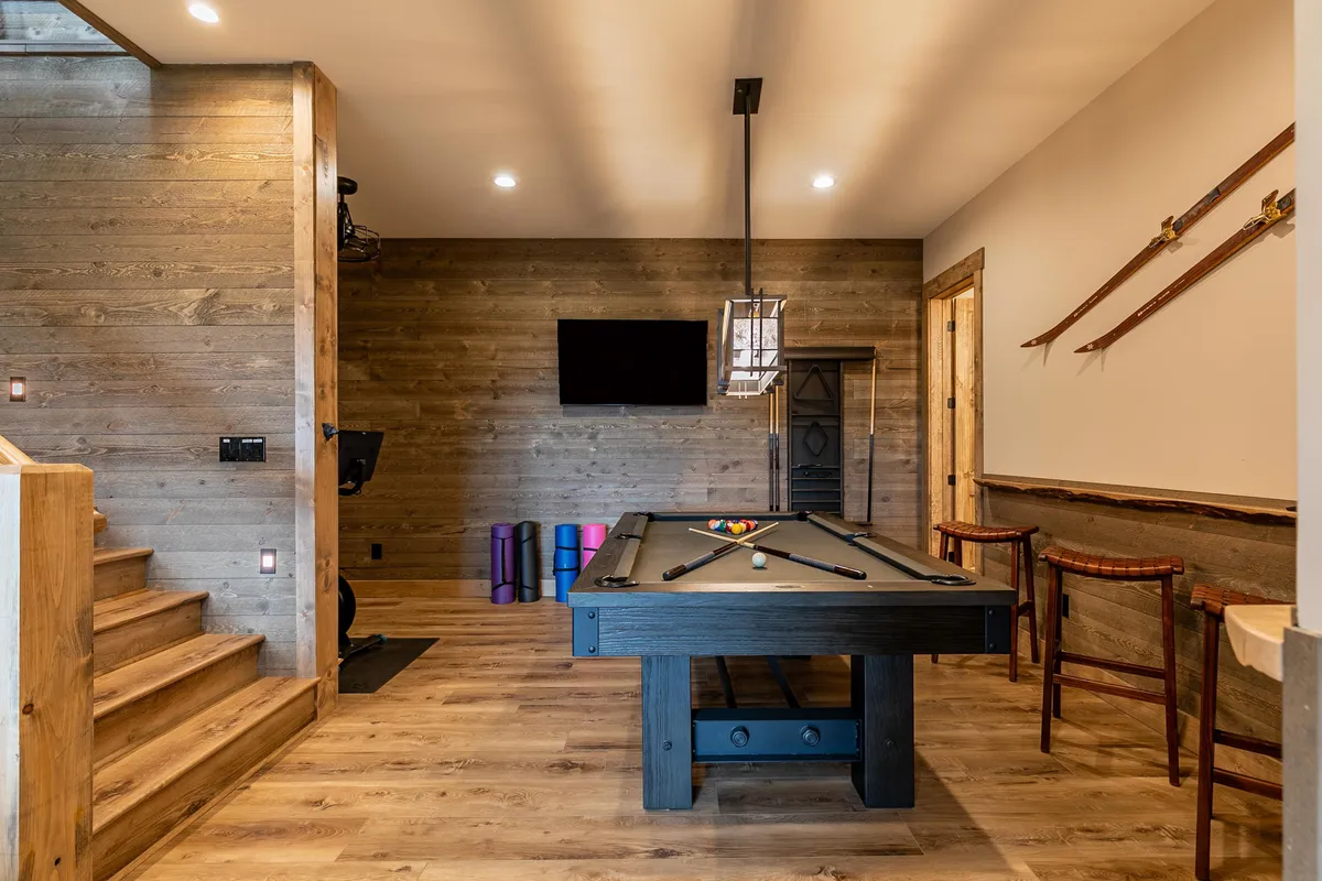 This image captures the recreation room’s central entertainment zone—a pool table, television, exercise equipment, and wooden stairs. The room’s layout blends classic games, entertainment, and fitness amenities, inviting both play and relaxation. The presence of swords on the wall adds a quirky, fun touch.