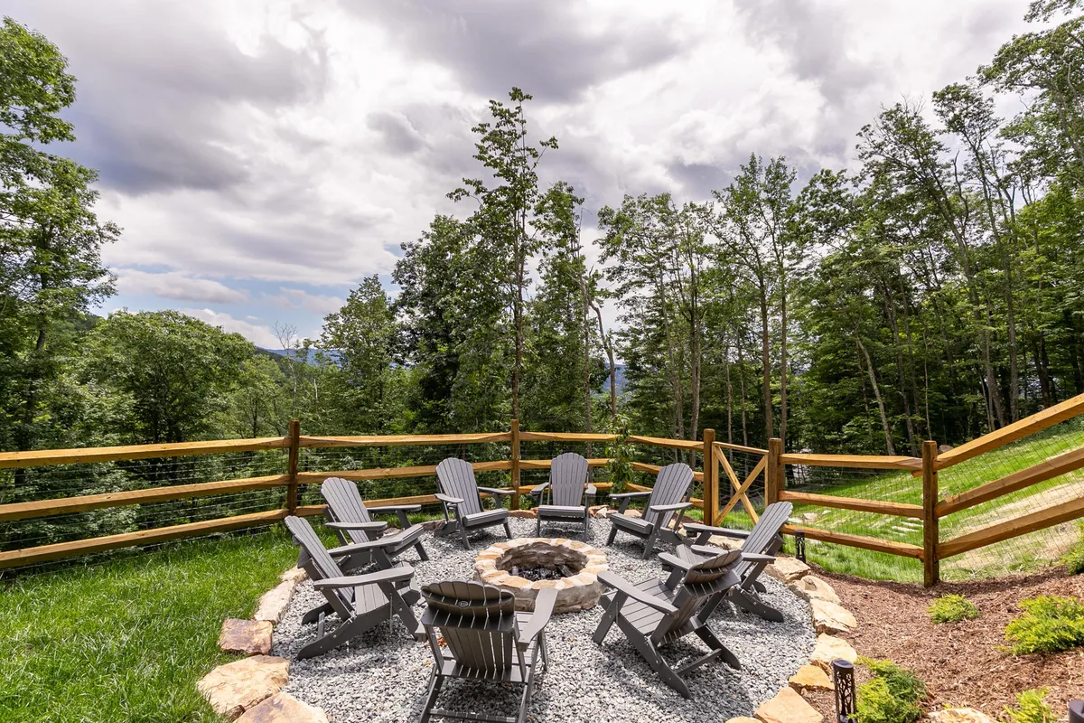 The photo shows a cozy fire pit area surrounded by adirondack chairs and natural landscaping, perfect for outdoor gatherings, storytelling, and relaxing with friends or family in the evening.