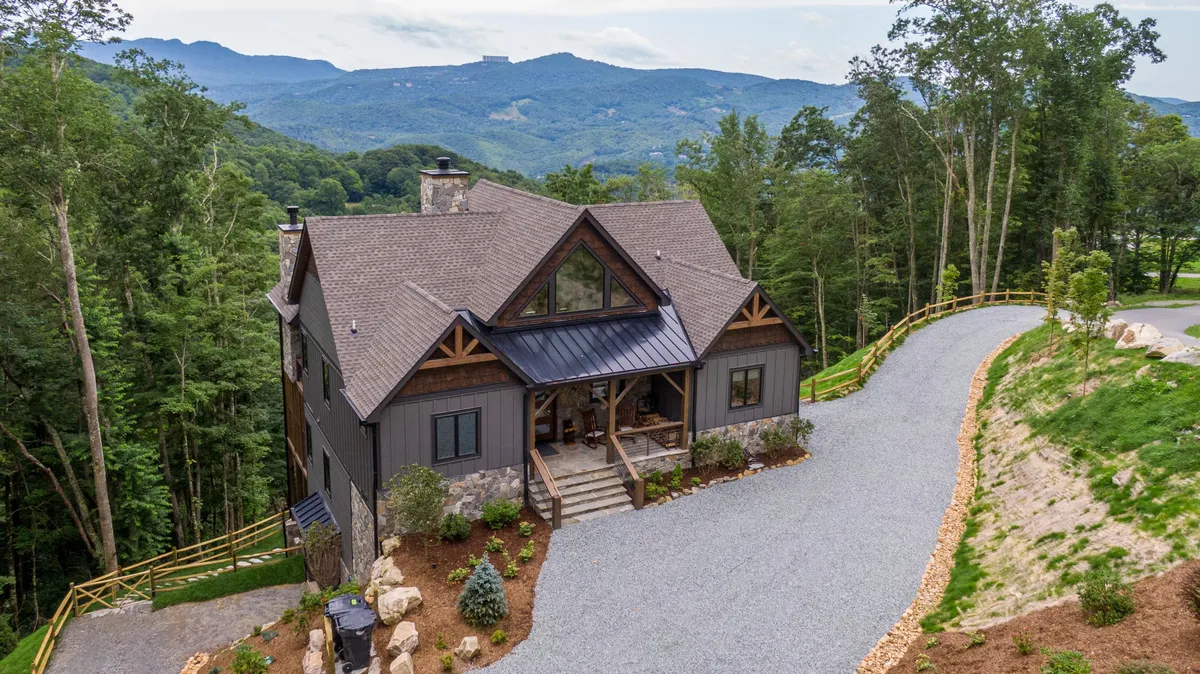 The image showcases the impressive facade of a luxurious mountain lodge, set against a stunning backdrop of Grandfather Mountain. A landscaped garden and spacious driveway lead up to the home, while a generous porch hints at sunrise views and space for relaxation. The serene mountain setting evokes a sense of tranquility and adventure.