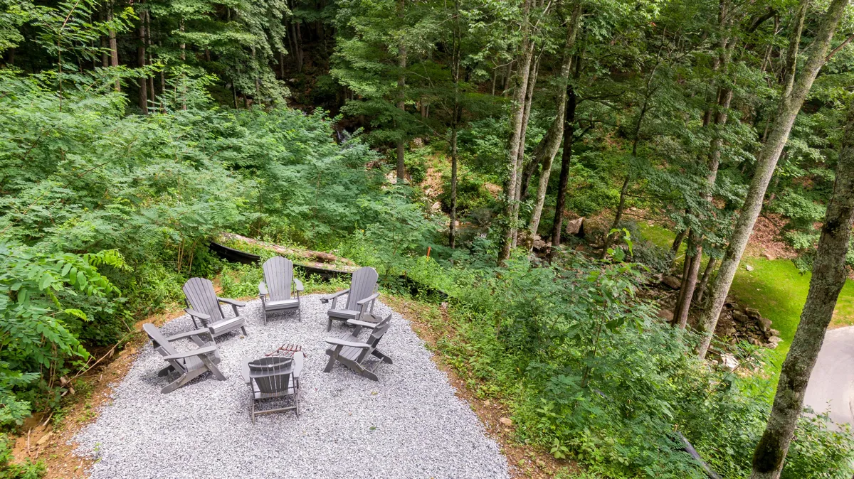 A gravel patio with a fire pit surrounded by Adirondack chairs, encircled by mature trees. The area evokes a sense of peaceful seclusion, ideal for evening relaxation, storytelling, or stargazing in a natural mountain setting.