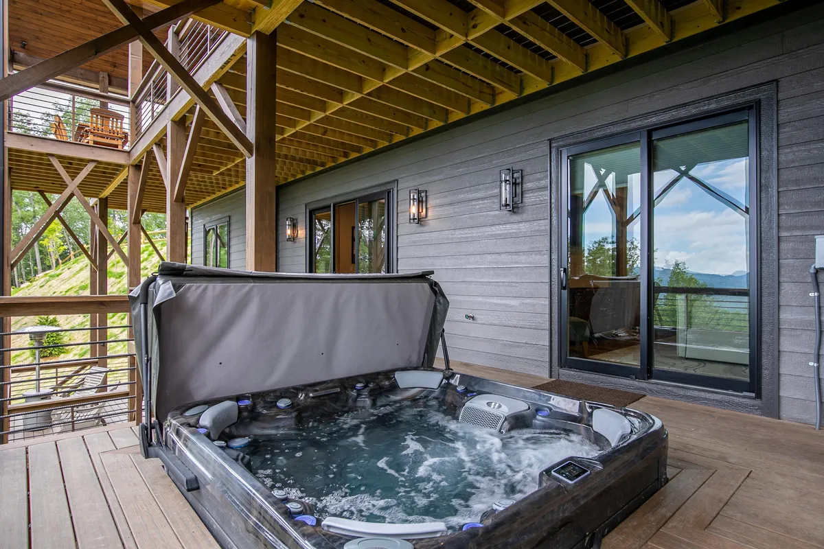 The image shows a serene outdoor deck featuring a bubbling hot tub, plenty of deck space, and a scenic view of the surrounding forest. The atmosphere is relaxed, offering multiple options for both private relaxation and socializing, such as stargazing or enjoying a morning coffee outside.