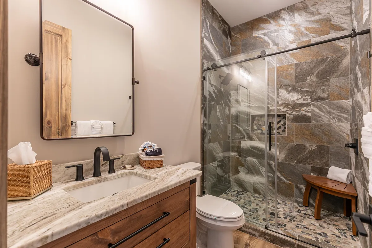 A contemporary bathroom with a granite countertop, a glass shower enclosure, wooden accents, and a large mirror. The clean lines and calming aesthetic support relaxation and self-care routines, echoing the pampered experience offered at Livin’ Lodge.