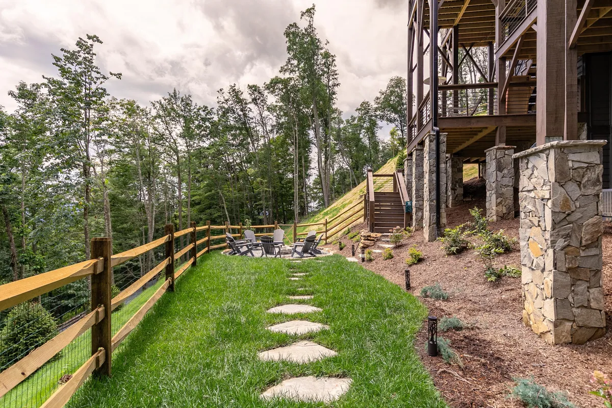 This exterior image highlights a spacious backyard featuring a stone walkway leading to a fire pit surrounded by trees and a deck attached to the house. The natural setting is ideal for outdoor gatherings, relaxation, and enjoying the peaceful mountain environment.