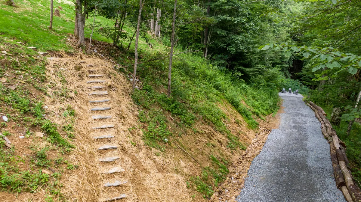 A gently ascending stone staircase and gravel path wind through lush greenery and mature trees, inviting exploration deeper into the natural landscape. The surrounding foliage hints at seasonal changes and a sense of adventure right outside the door.