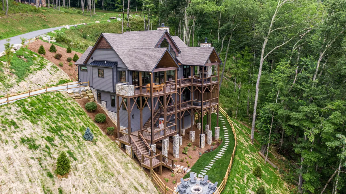 This exterior image shows the elevated structure of Livin' Lodge at Eagles Nest, featuring stone pillars, green landscaping, and a central fire pit area surrounded by comfortable seating. The deck extends out, inviting guests to enjoy the surrounding nature.