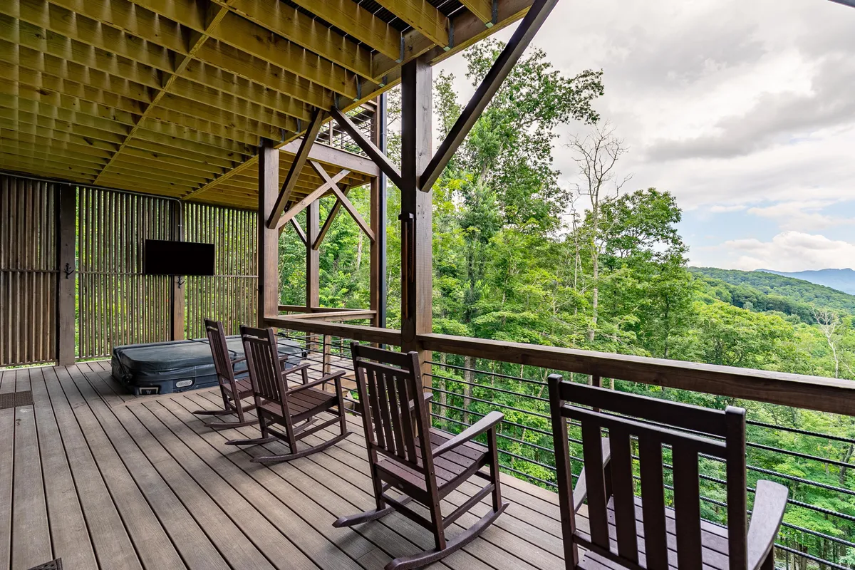 The exterior deck offers a serene retreat with a hot tub overlooking trees and mountains, wooden rocking chairs, and a welcoming railing. The scene radiates peace, inviting guests to relax, enjoy nature, or stargaze from the bubbling hot tub.