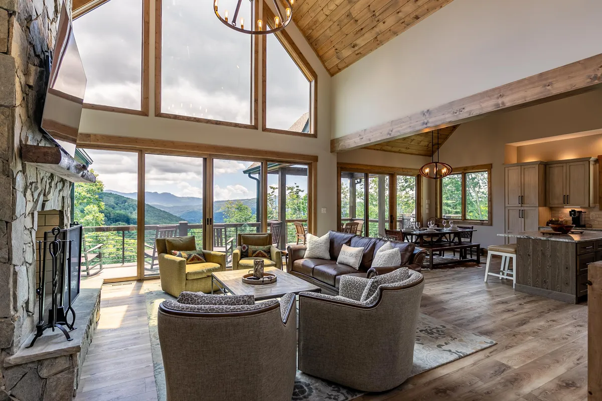 This image captures a grand Great Room with soaring windows that frame stunning mountain views. The space features a prominent stone fireplace, comfortable seating, and an adjacent dining area, all bathed in natural light. The open layout and mountain vistas encapsulate the central gathering space described in the section.