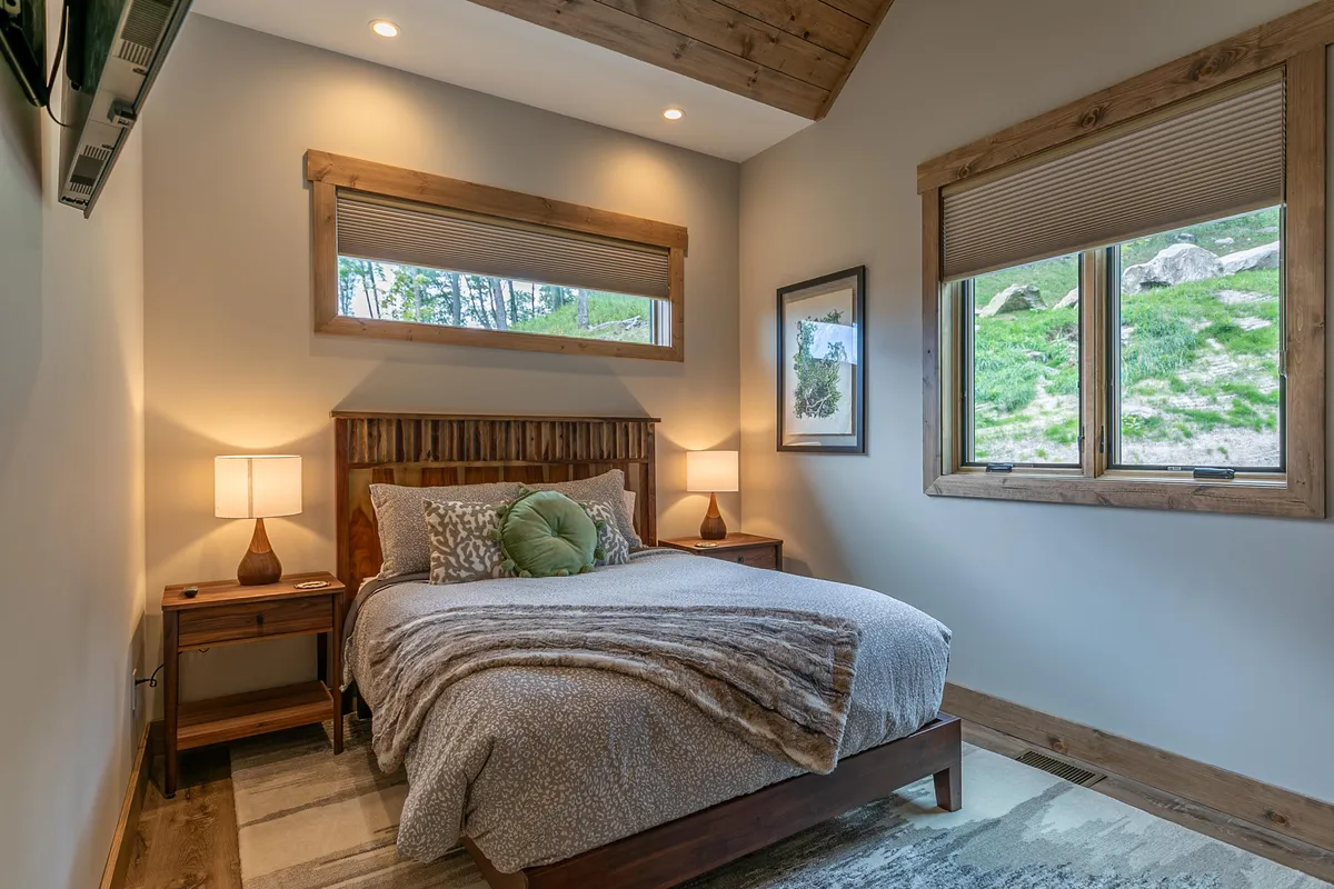A tranquil bedroom featuring a plush bed adorned with decorative pillows, wooden nightstands with soft lamps, framed artwork on the wall, and expansive windows that frame vibrant greenery outside. The room is flooded with natural light, creating a peaceful and restful ambiance ideal for restorative sleep or a quiet morning in bed.