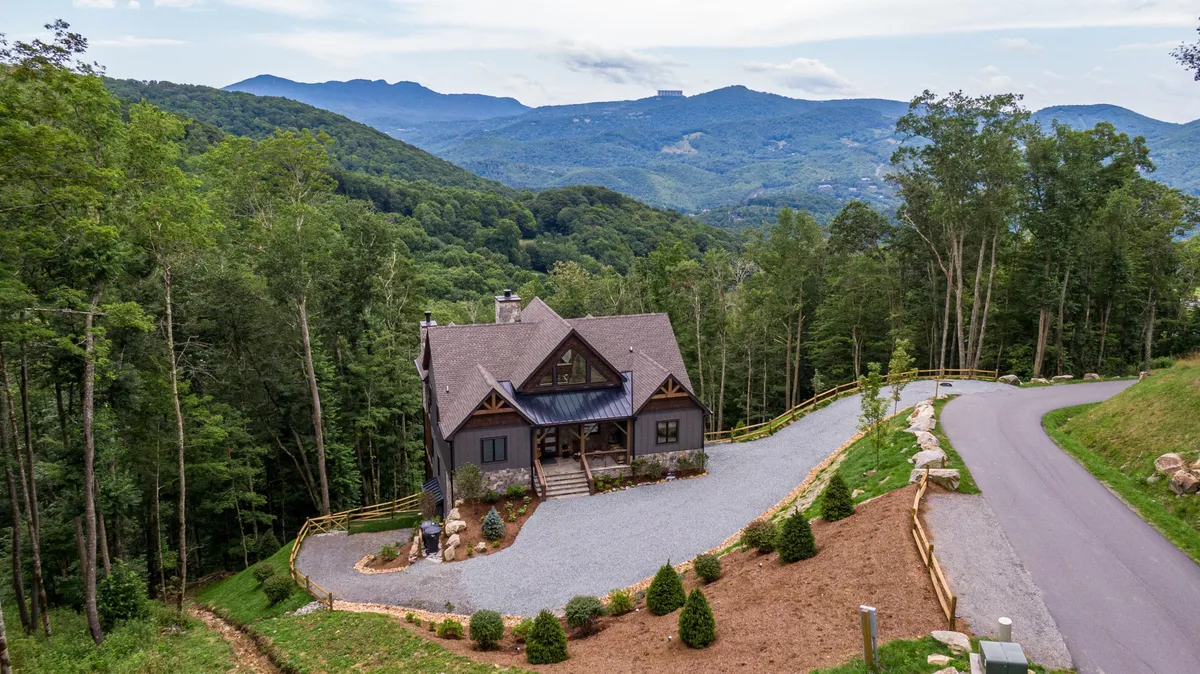 This exterior shot presents the main house nestled among trees with mountain views in the distance. The driveway and porch are visible, underscoring easy access and the lodge's integration into the natural setting. The sense of arrival and the broader resort community context are both conveyed here.