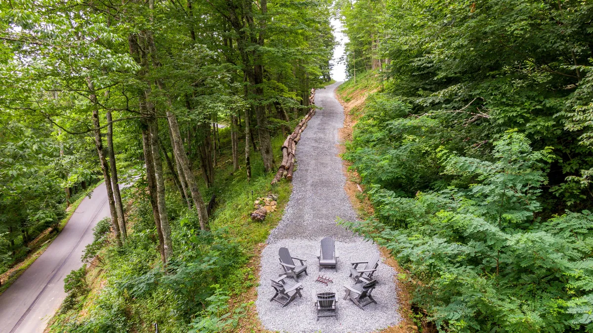 An intimate outdoor scene with a gravel path, cozy seating circling a fire pit, and tall trees providing shade and privacy. The layout invites small groups or individuals—perhaps with their dogs—to gather, share stories, or relax in nature.