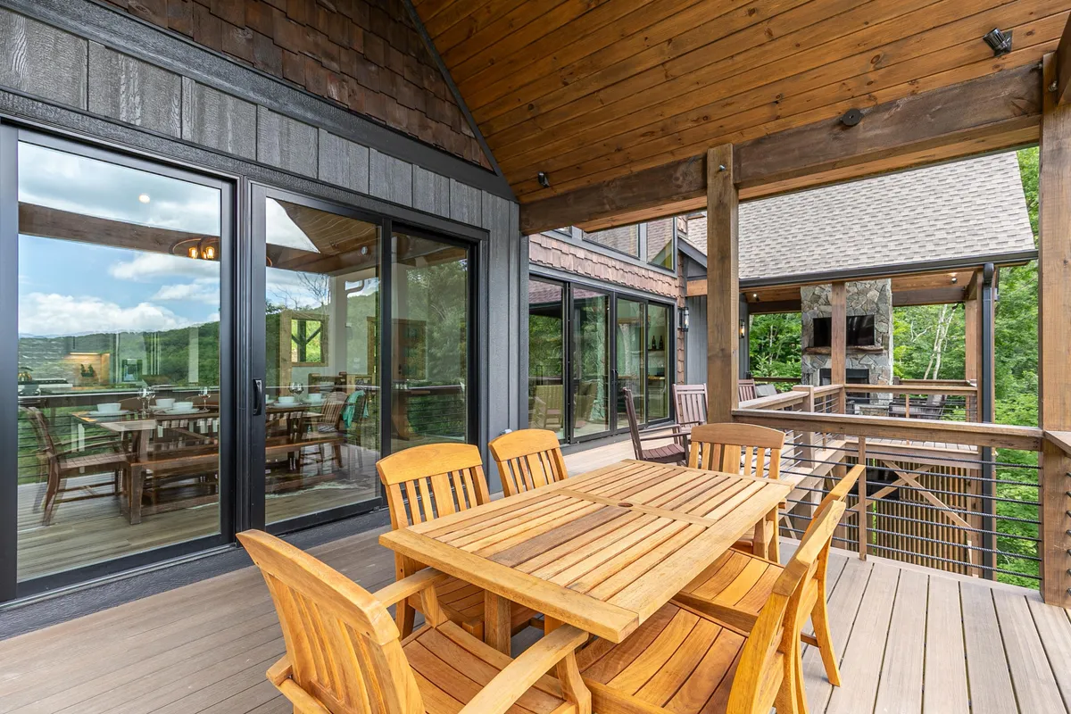 An inviting outdoor dining area featuring a wooden table set amidst lush greenery and large glass doors that connect seamlessly to the indoors. The setting is ideal for outdoor meals, gatherings, and social occasions in a tranquil natural environment.
