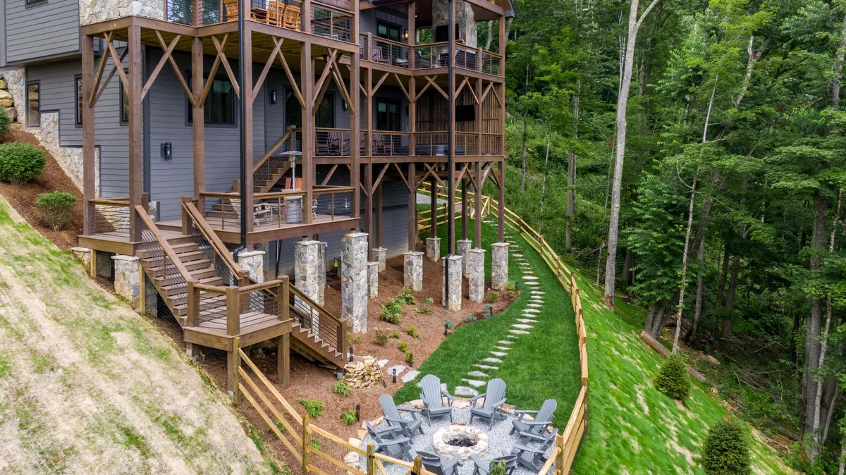 This image captures a spacious multi-level wooden deck surrounded by trees, complete with a cozy fire pit area and sturdy stone pillars. The setup is perfect for groups to gather outdoors for dining, storytelling, or enjoying nature together. The lush greenery and layered deck design highlight accessibility and a welcoming communal atmosphere.